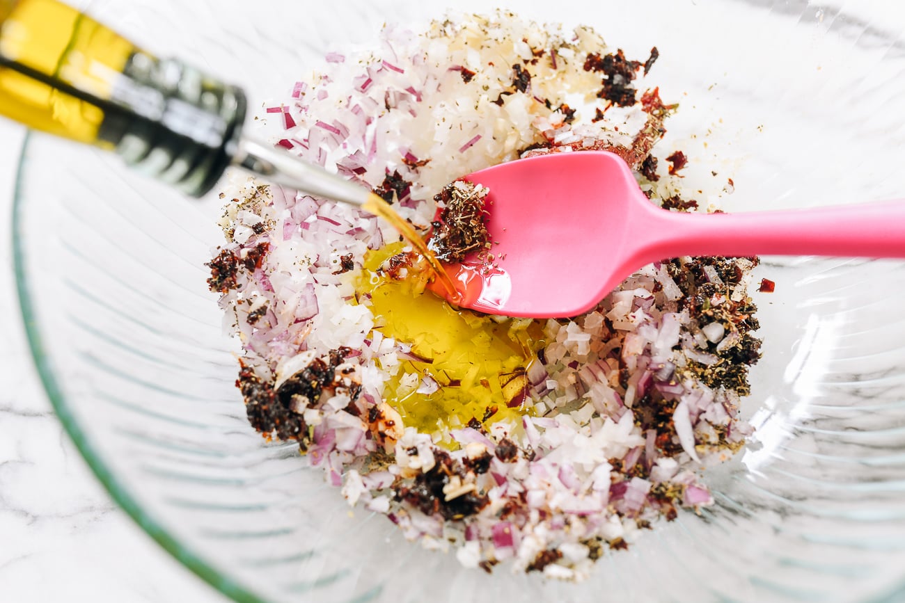 adding olive oil to onion, garlic, herb mixture in glass bowl