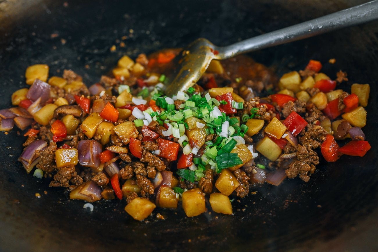 wok spatula stirring braised ground pork and potato in wok with green scallions over top