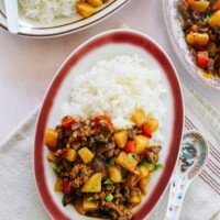 Chines braised pork, potato, and bell pepper over rice on a red rimmed oval plate on a pink marble counter
