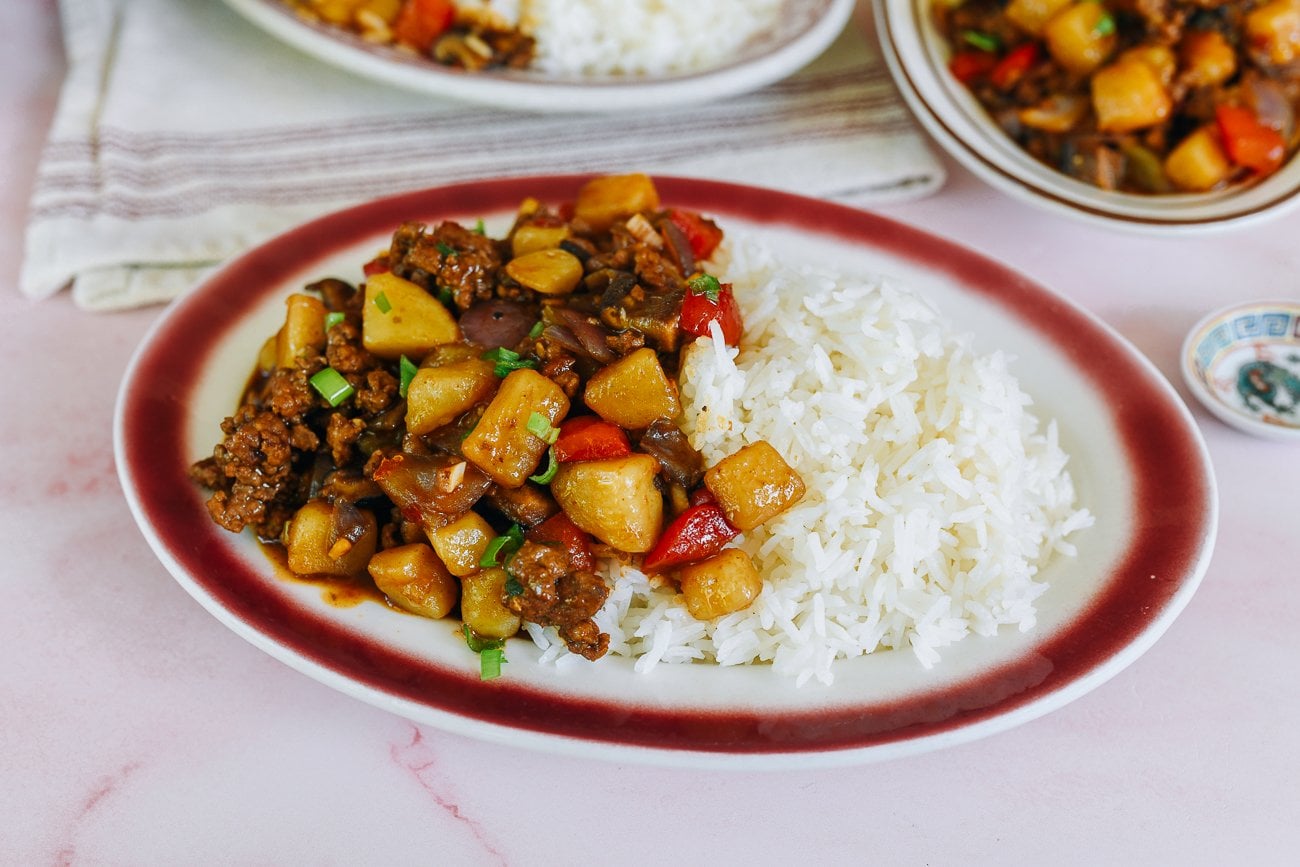 Chines braised pork, potato, and bell pepper over rice on a red rimmed oval plate on a pink marble counter