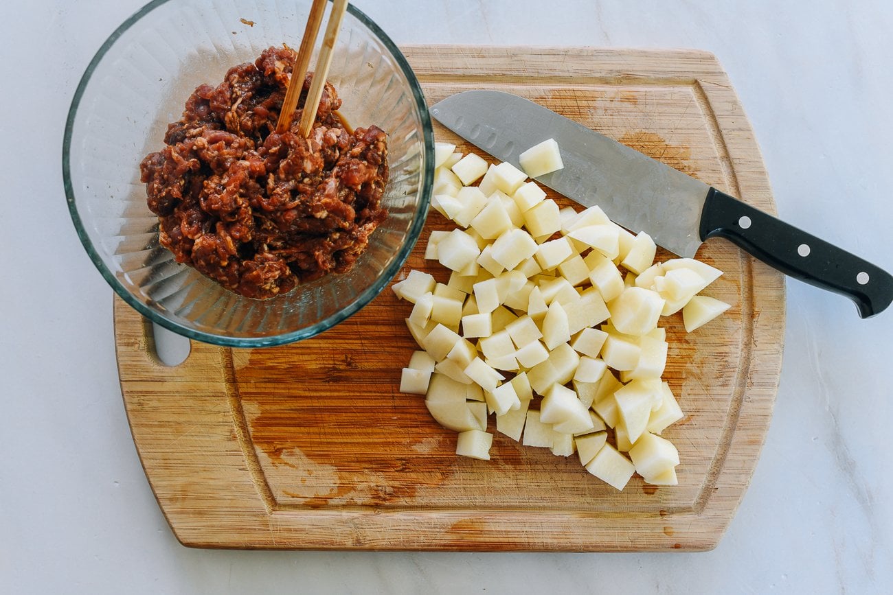 Clear bowl of minced soy sauce marinated pork and cubed potatoes on a cutting board