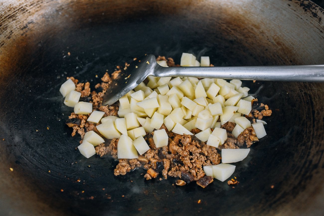 Cubed potatoes over pork and mushroom mixture in a wok with wok spatula