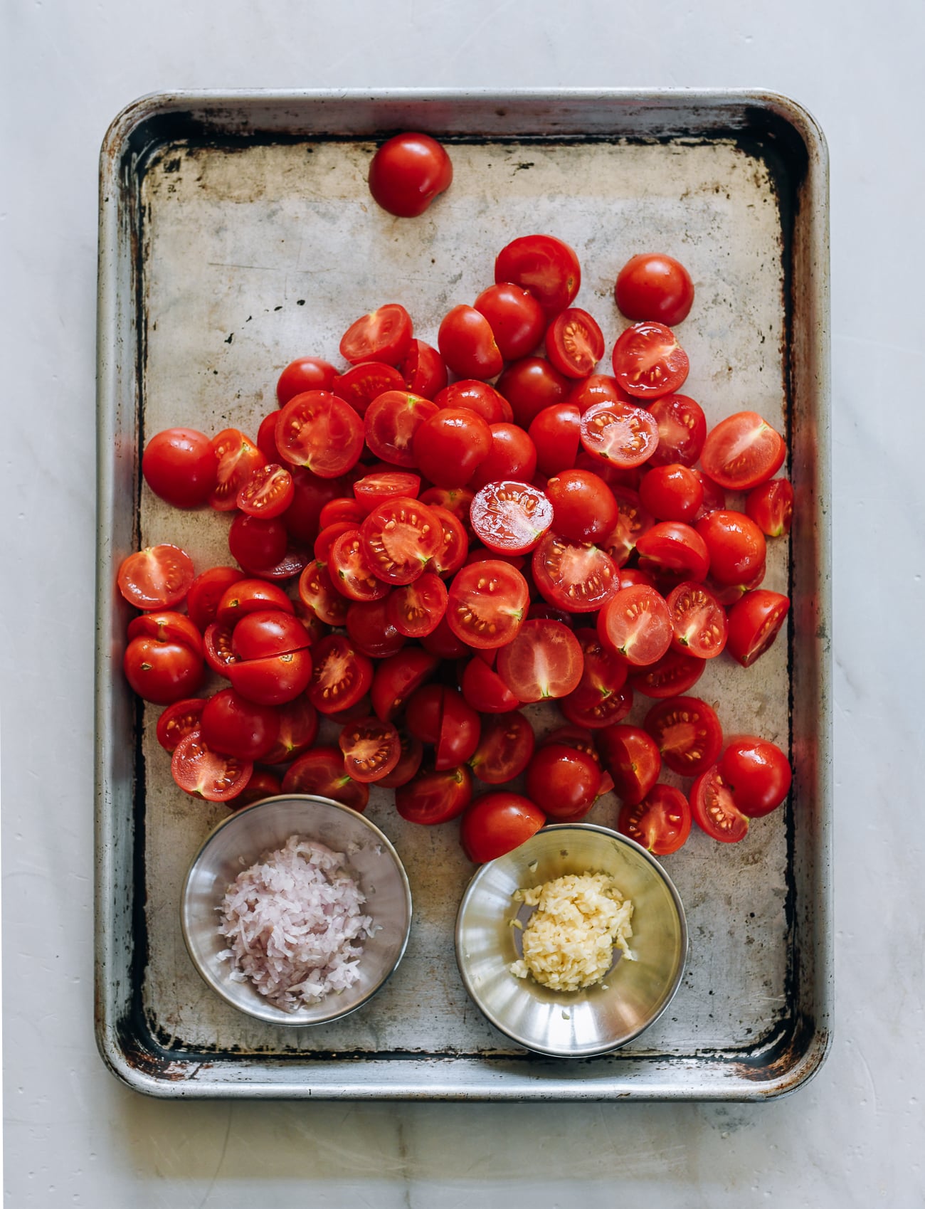 Tomato halves on sheet pan with two pinch bowls of chopped shallot and chopped garlic