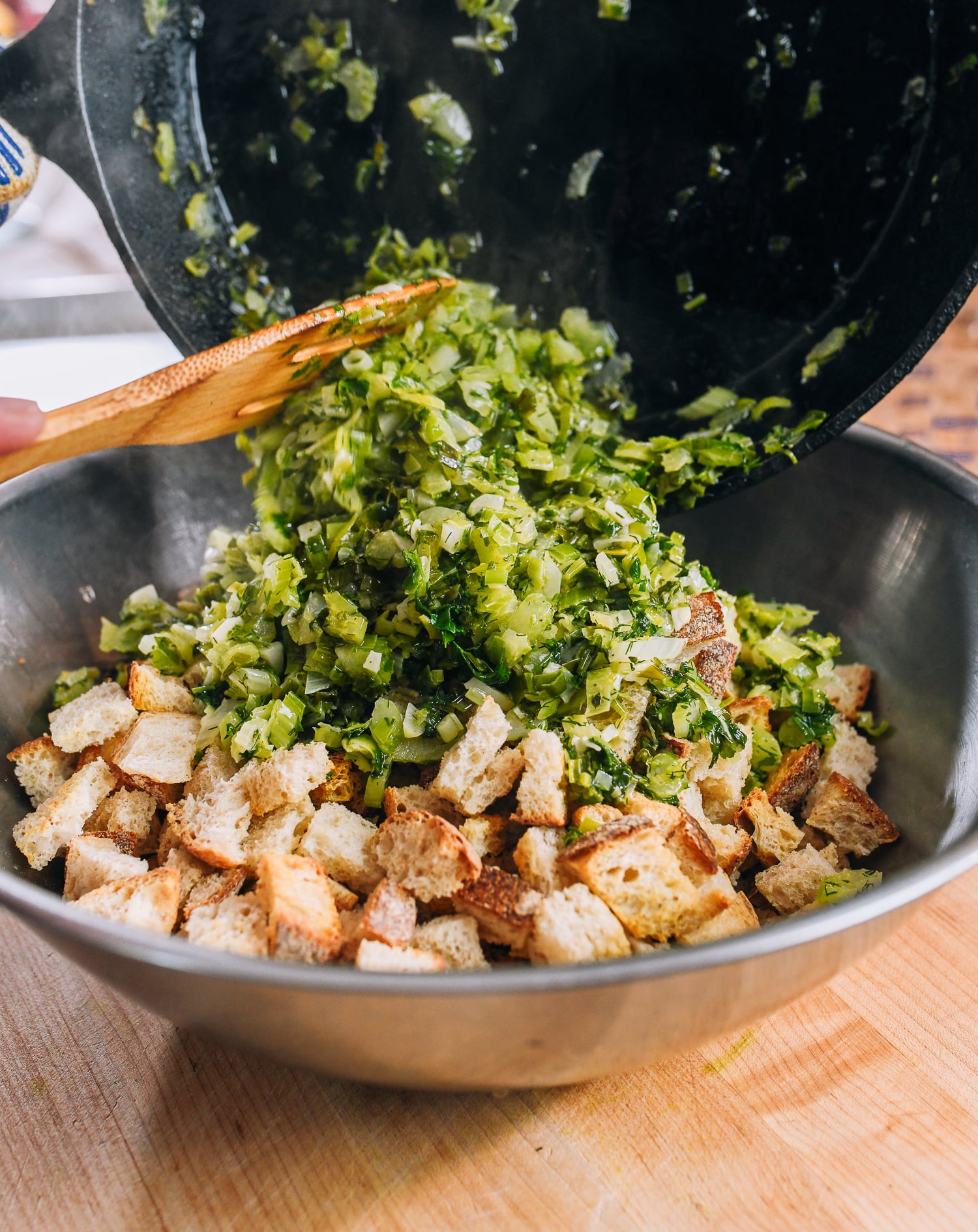 adding vegetable and herb mixture to bread cubes in stainless steel bowl
